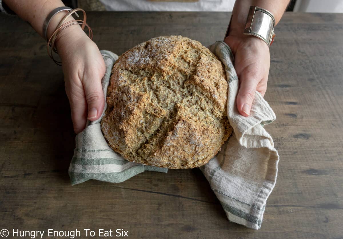 Hands holding a round brown bread loaf in a dish towel.