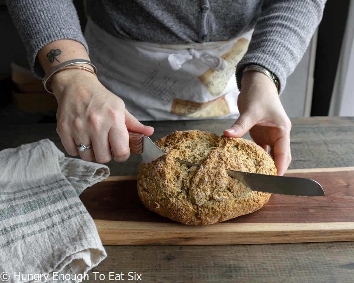 Hands holding and slicing loaf of brown soda bread.
