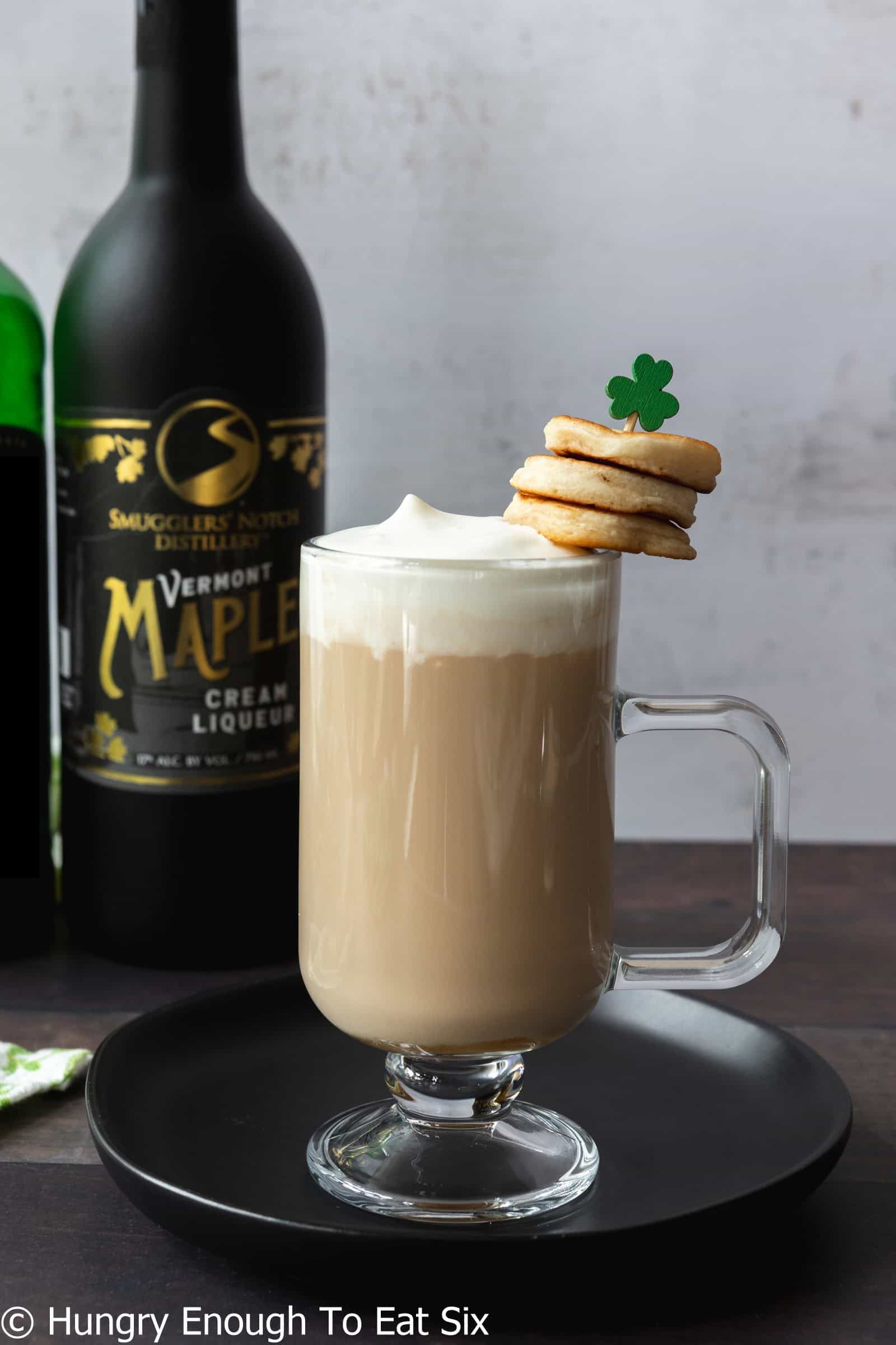Glass mug of Irish coffee with liquor bottles behind it.