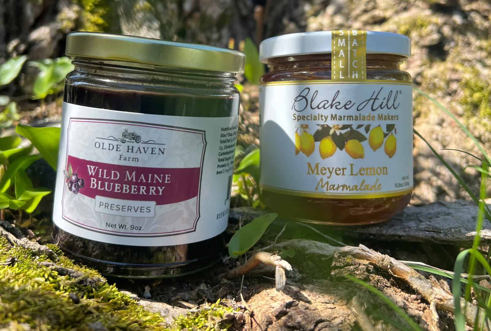 Two brands of jarred jam and marmalade on an outdoor grassy spot.